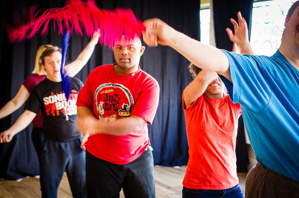 A group of people dancing indoors, some holding colourful streamers. They appear to be exercising or participating in a community dance class or workshop, with focused and energetic expressions. Black curtains are in the background.