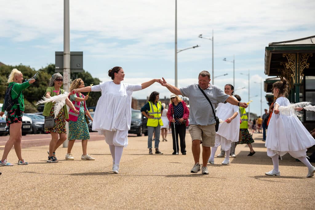 A man joins a lively dance with a performer in white, surrounded by other dancers and festival-goers on a sunny promenade. Onlookers take photos, while some people in high-visibility vests help keep the community event running smoothly.